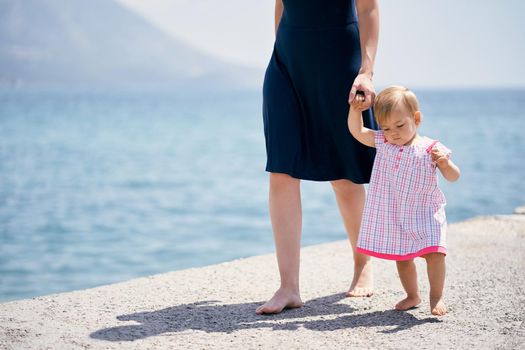 Mom leads a little girl by the hand along a concrete breakwater. High quality photo