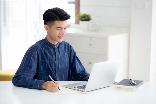 Young asian business man writing on notebook for planning working and using laptop computer on desk at home, notes about finance, male study and learning, business and communication concept.