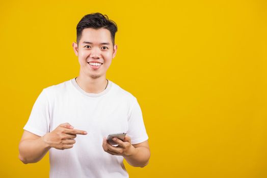 Asian handsome young man smiling positive holding smartphone blank screen and pointing finger to mobile phone, studio shot isolated on yellow background, making successful expression gesture concept