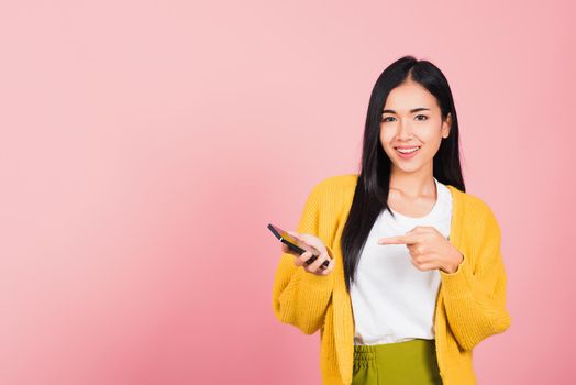 Portrait Asian happy beautiful young woman smile making finger pointing on smartphone in hand shopping online application looking to camera isolated, studio shot pink background with copy space