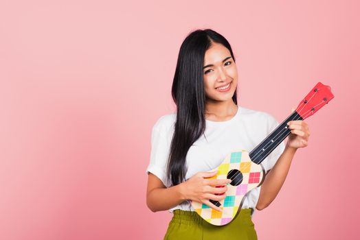 Portrait of happy Asian beautiful young woman teen confident smiling face hold acoustic Ukulele guitar, female playing Hawaiian small guitar, studio shot isolated on pink background, with copy space