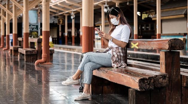 Asian tourist teenage girl wearing mask waiting distance train station using smartphone, social media check-in, lone traveler, Summer vacation railroad adventure concept