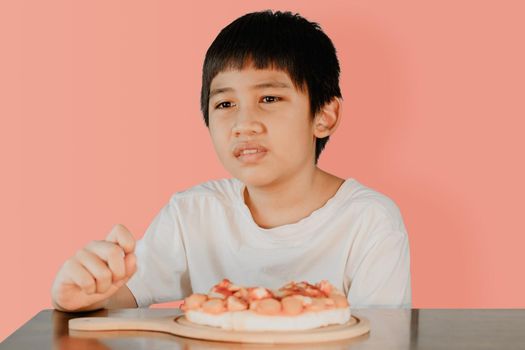 Asian cute boy sitting at the dining table with pizza on the table in front on pink color background.