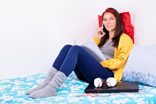 Beautiful young girl sitting on her bed in her room holding a book and talking on a mobile phone. Young girl in bed smiling and talking on the phone. Communication, friendship, quarantine concept.