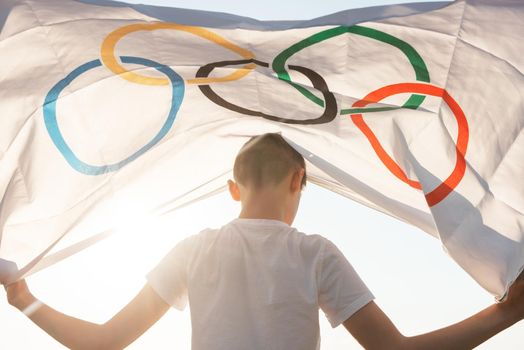 Portrait of boy waving flag the Olympic Games outdoors over cloudy sunset sky. Children sports fan. Summer olympic games concept. 08.06.2021, Barnaul Russia.