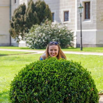 A young beautiful girl looks out from behind a spherical bush. Hiding from friends.