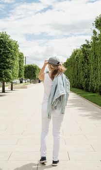 Woman in casual jeans clothes walking in the park. Back view of a blond woman holding her cap walking outdoors