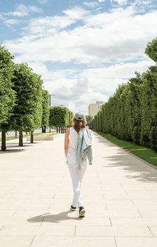 Woman in casual jeans clothes walking in the park. Back view of a blond woman holding her cap walking outdoors