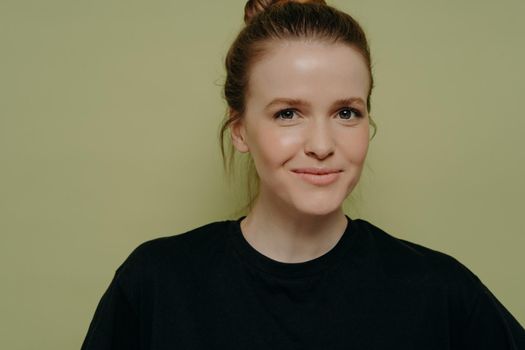 Close up portrait of young cute female stud t in black tee shirt with hair in bun, looking straight at camera with slight smirk, trying to hold her laugh, standing isolated on light green background