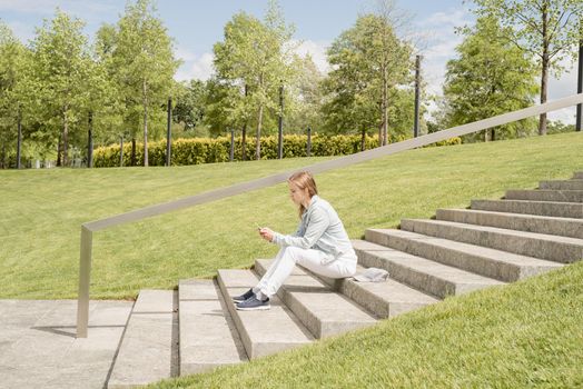 Woman in jeans clothes texting on her mobile phone, sitting on the stairs in the park