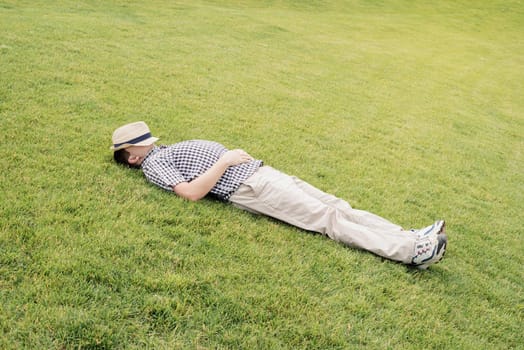 Young thoughtful man in summer hat sitting on the grass in the park, looking away