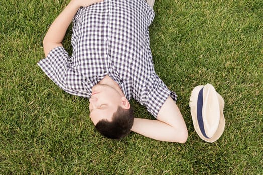 Young thoughtful man in summer hat sitting on the grass in the park, looking away