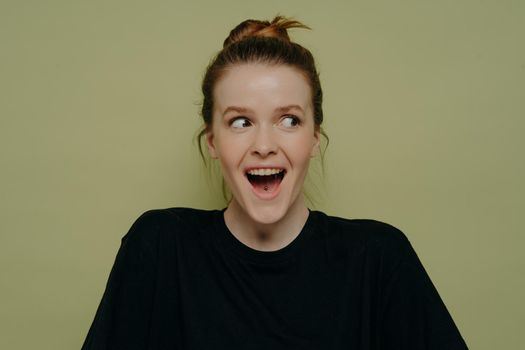 Young amazed woman in casual outfit being surprised after hearing amazing story or cant believe her eyes, looking aside with open mouth while seeing something amazing, isolated on green background