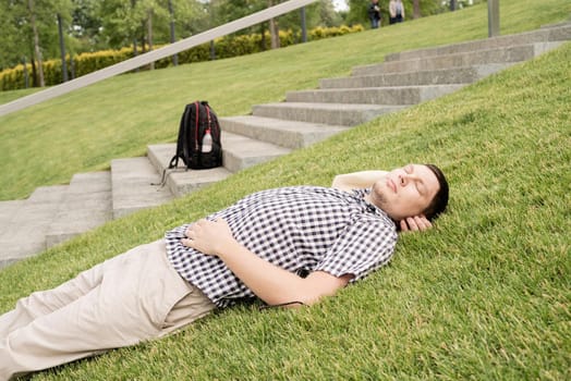 Young thoughtful man in summer hat sitting on the grass in the park, looking away