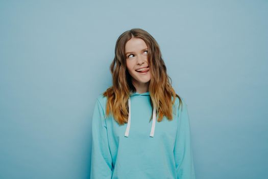 Young carefree pretty teenage girl with amazing long wavy light hair, dressed in casual light blue hoodie, making crazy funny expression with amazing smile showing her tongue alone on blue background