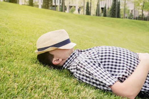 Young thoughtful man in summer hat sitting on the grass in the park, looking away