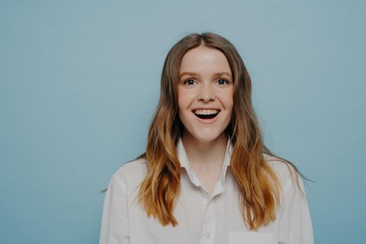 Happy young woman dressed in white shirt surprised after being informed about getting office position at company afterwards job audition, smiling and feeling excited while posing next to light wall