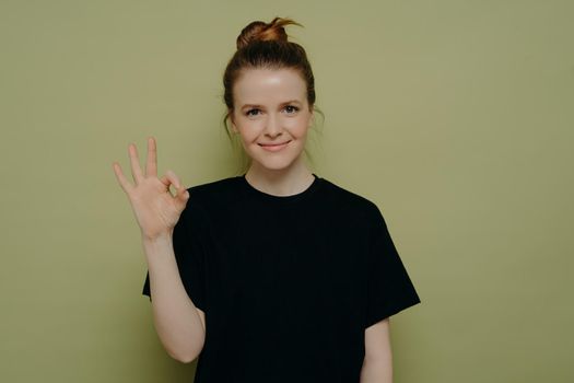 Calm brunette woman in black tee shirt with hair in bun and slight smile, making okay sign with her hand and feeling satisfied, showing that everything is awesome, isolated in front of green wall
