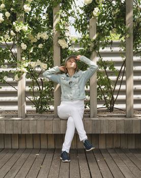 Woman in casual jeans sitting on the wooden bench in green park alley, walls with white roses. Light and shadow pattern