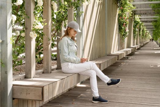 Woman in casual jeans clothes sitting on the wooden bench using phone in green park alley, walls with white roses