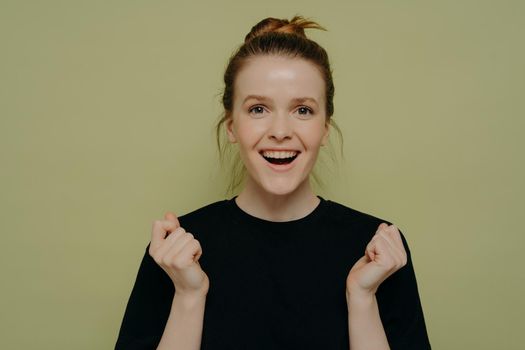 Cheering happy teenage girl in black shirt with hair in bun overwhelmed with positive emotions and can not believe her eyes, laughing with fists in air while standing isolated on green background