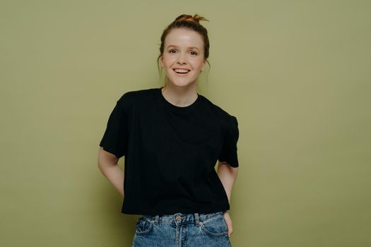 Young happy young woman with hair in bun dressed in casual clothes standing in relaxed posture and looking at camera with wide smile on face while posing in studio on green background