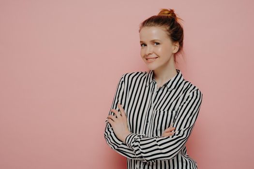 Adorable happy ginger teenage girl in striped shirt with hair in bun looking at camera with bright smile and expressing positive emotions, standing sideways with crossed arms against pink background