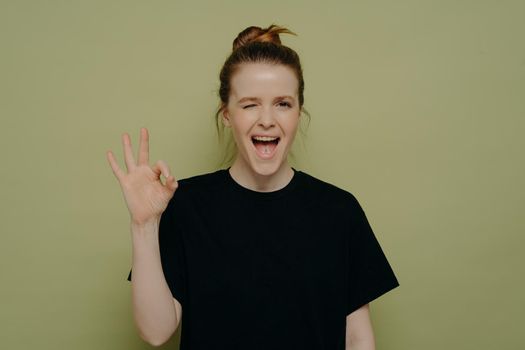 Overjoyed excited young woman in black tee shirt winking with open mouth, making ok sign with hand, assuring everything is going good and well, posing against green wall