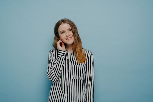 Tender cute european teen girl wearing white shirt with black stripes, expressing her shyness with tilted head and hair behind ear, posing in studio in front of light blue background