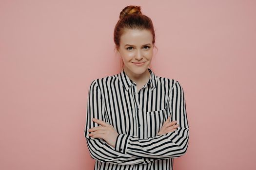 Satisfied happy redhead teen girl with hair in bun looking straight and smiling joyfully at camera with arms crossed, isolated in front of pink background, waist up portrait of young delighted woman