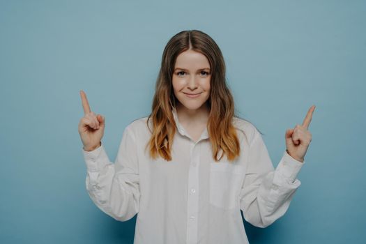 Beautiful teenage girl in white shirt with smug smile and satisfied emotion on face pointing up with her fingers, indicating at copy space, isolated over blue background. Sale and promotion concept