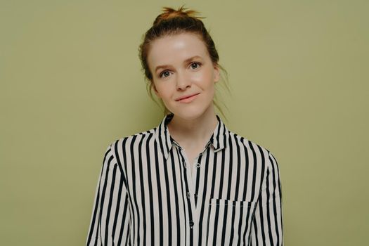 Portrait of calm young european woman with hair in bun wearing black and white striped shirt looking at camera with tilted head and slight smile while standing alone next to green wall