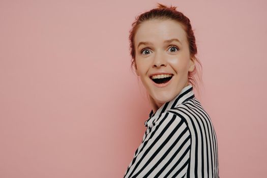 Greatly surpirsed redhead teenage girl with hair in bun, wearing white striped shirt, turning her head back, shocked by unexpected news, standing isolated next to pink wall. Human emotions concept