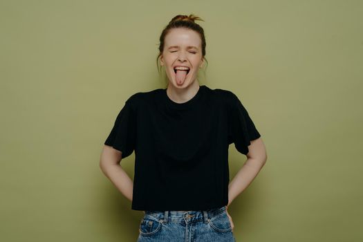 Crazy young woman wearing casual clothes with hair in bun making a goofy face with wide open mouth, showing her tongue and having fun while standing isolated next to green background