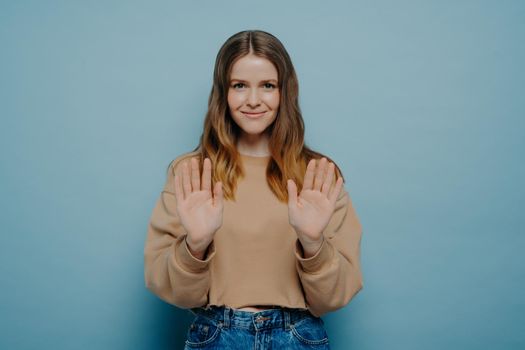 Young woman in beige sweater expressing disagreement, showing it by holding hands in front of her, saying no with her gesture, politely refusing to do what she asked on light blue background