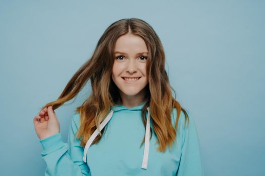 Cute cheering expressive teenage girl with smiling expression in blue hoodie playing with her long wavy hair while looking at camera with positive expression isolated on light blue background