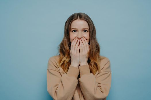 Young excited lady in beige sweater looking forward as she is surprised by event or news, holding hands against mouth and looking at camera with shocked face expression, isolated on blue background