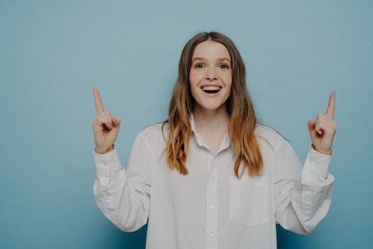 Amazed young woman pointing upwards with fingers and expressing excitement while seeing something amazing, isolated on blue wall, offering great opportunity. Advertisement and promotion concept