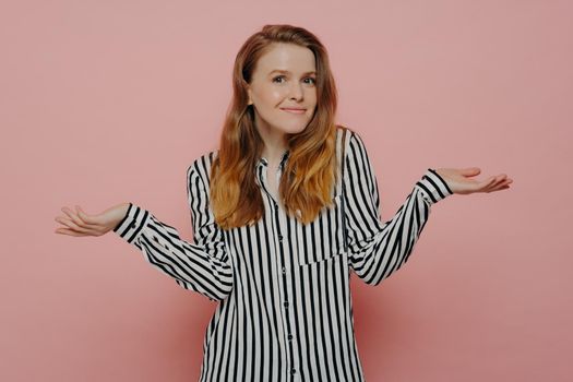 Young doubtful female dressed in white shirt with black stripes, shrugging with both hands in air, doesnt know what to do while stnading next to pink wall in studio. Teenage girl looking uncertain