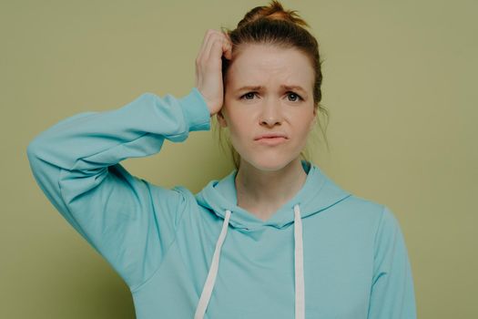 Young puzzled female student in blue hoodie expressing confused emotion, holding hand on head and being frustrated by situation, standing isolated on green background in studio
