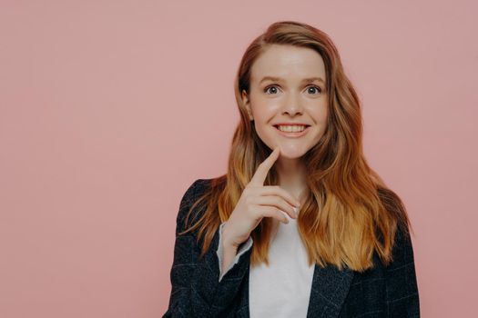 Young excited ginger lady in jacket with white shirt, realizing there is solution, generating new idea with finger on chin, standing alone next to pink background and expressing happiness