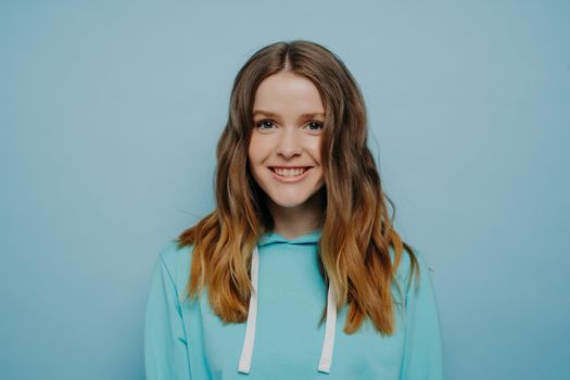 Long haired young happy attractive woman dressed in urban style hoodie standing against blue studio background and smiling, headshot of cheerful teenager being in good mood