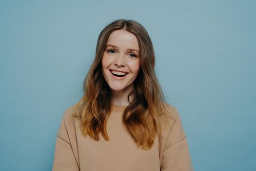 Portrait of cute laughing european teenage girl dressed in casual clothes smiling at camera with positive face expression while standing isolated blue background. Happy people concept