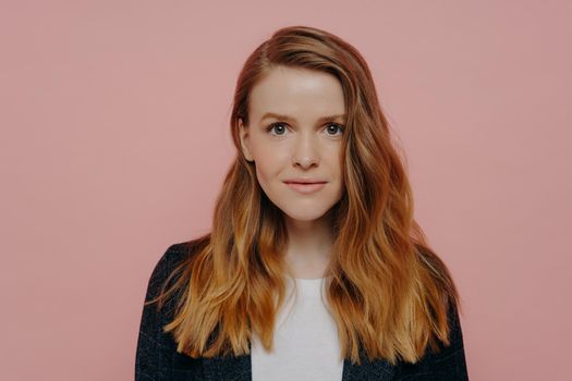 Portrait of smiling young redhead woman with wavy hair wearing casual clothes looking at camera confidently, posing on color background in studio, female natural beauty concept
