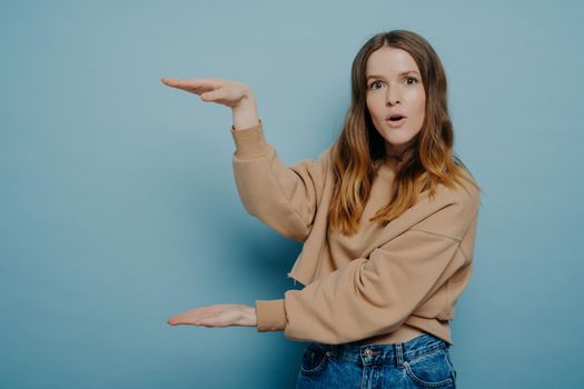 Surprised young woman wearing casual clothes presenting size of product with wow face expression while standing isolated over blue background, female showing big large size with hands