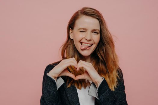 Attractive carefree young woman with wavy ginger hair in formal dark jacket showing heart shape sign, winking and sticking tongue out standing against light pink wall in studio, expressing fun