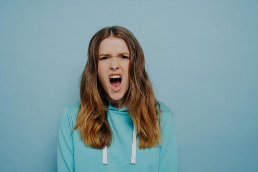 Emotional teenage girl expressing anger and disbelief while looking at camera with open mouth and eyebrows pulled close together, standing against blue background. Negative human emotions concept