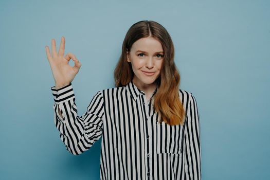 Smiling confident young woman with wavy brown hair showing okay sign with one hand wearing stripy black and white blouse while posing isolated over blue light blue studio background