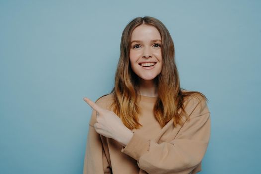 Smiling pretty young female with balayage hair in loose casual sweater pointing her finger up at something attracting attention, standing against blue wall in studio. Advertisement and promotion