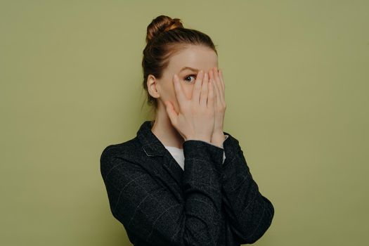 Photo of young ginger woman with bun closing face with both hands showing one eye looking at camera with fear, wearing dark formal jacket and white top posing against light grey wall